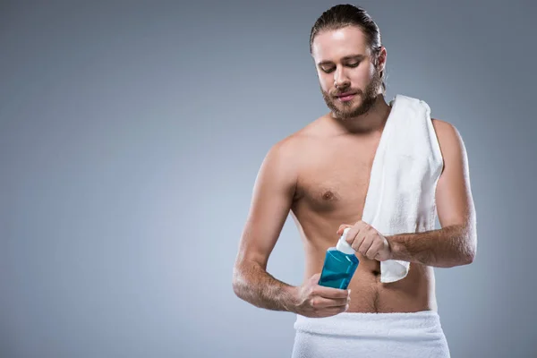 Young man with bath towel on his shoulder holding bottle with tooth rinse in hands,  isolated on gray — Stock Photo