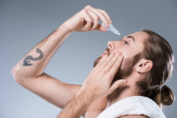 Portrait of handsome young man dripping eye drops with hand on cheek, isolated on gray — Stock Photo
