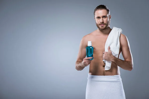 Mann mit Badetuch auf der Schulter hält Flasche mit Zahnspülung in der Hand, isoliert auf grau — Stockfoto