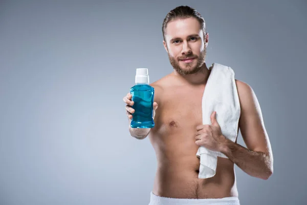 Smiling man with bath towel on shoulder holding bottle with tooth rinse in outstretched hand, isolated on gray — Stock Photo