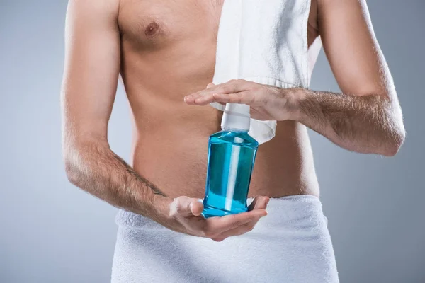 Midsection of man with bath towel on shoulder and around waist holding bottle of tooth rinse in hands, isolated on gray — Stock Photo