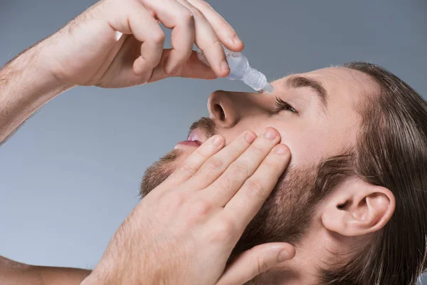 Portrait of handsome young man dripping eye drops with hand on cheek, isolated on gray — Stock Photo