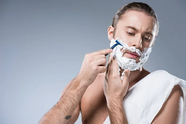 Homem caucasiano jovem com espuma de barbear no rosto segurando navalha na mão contra sua bochecha, enquanto toalha de banho que coloca em seu ombro, isolado em cinza — Fotografia de Stock