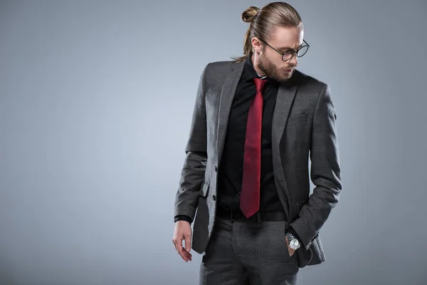 Homem de negócios morena de cabelos longos em óculos olhando para baixo com a mão no bolso, isolado em cinza — Fotografia de Stock