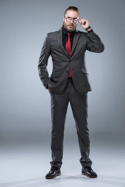 Бізнесмен в офіційному костюмі торкається окулярів, стоячи, ізольований на сірому — Stock Photo