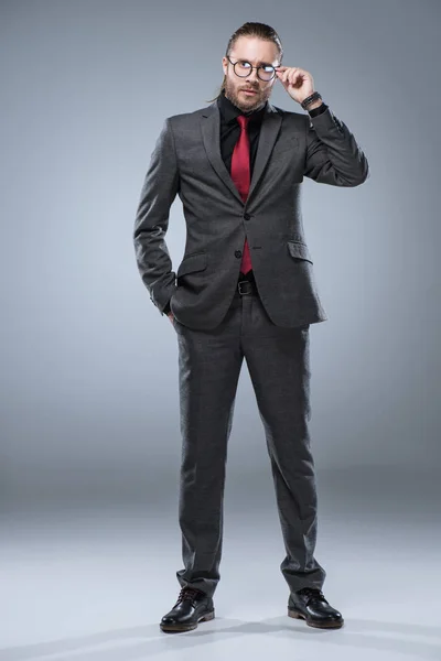 Businessman in formal suit touching his glasses while standing, isolated on gray — Stock Photo