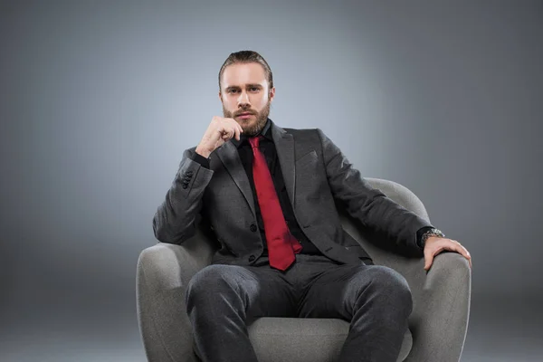 Sérieux jeune homme d'affaires en costume formel assis sur un fauteuil, avec la main sur le menton, isolé sur gris — Photo de stock