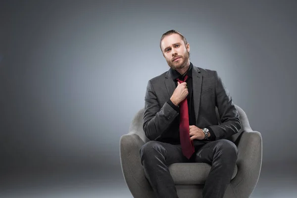 Homme d'affaires sérieux assis dans un fauteuil et redressant cravate, tout en regardant la caméra, isolé sur gris — Photo de stock