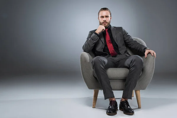 Серьезный молодой бизнесмен, сидящий в кресле с рукой на подбородке и смотрящий в камеру, изолированный на сером — стоковое фото
