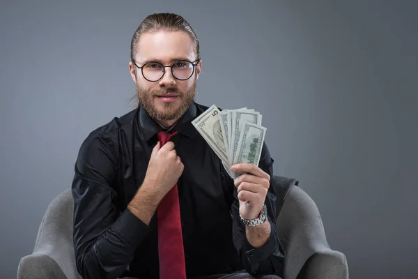 Erfolgreicher lächelnder Mann mit Geld in der Hand, der im Sessel sitzt, während er Krawatte richtet, isoliert auf grau — Stockfoto