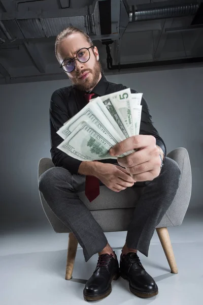 Seriöser Geschäftsmann mit Brille sitzt im Sessel, Geld in der Hand und blickt in die Kamera — Stockfoto