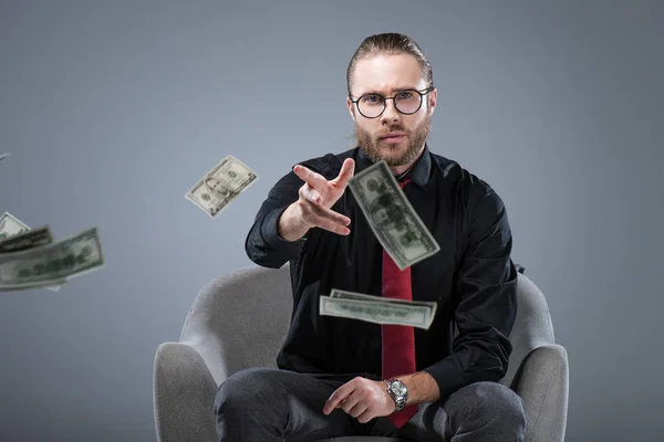 Giovane uomo d'affari di successo in occhiali seduto in poltrona mentre gettava soldi alla macchina fotografica, isolato su grigio — Foto stock