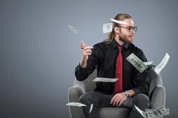 Empresario sentado en sillón mientras tira dinero, aislado en gris - foto de stock
