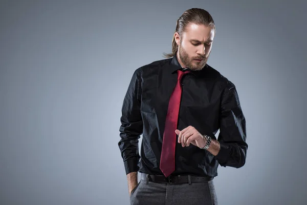 Серйозний чоловік в чорній сорочці з червоною краваткою дивиться на наручний годинник, ізольований на сірому — стокове фото