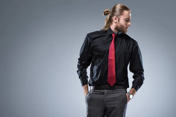 Joven hombre barbudo con camisa negra con corbata roja mirando hacia otro lado, aislado en gris - foto de stock