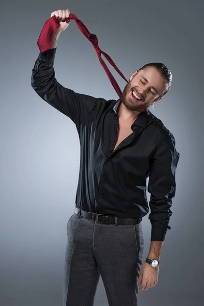 Smiling bearded man in black shirt holding red tie around his neck, isolated on gray — Stock Photo