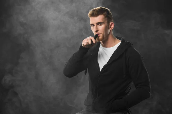 Молодой бородатый мужчина выкуривает электронную сигарету в окружении облаков пара — стоковое фото
