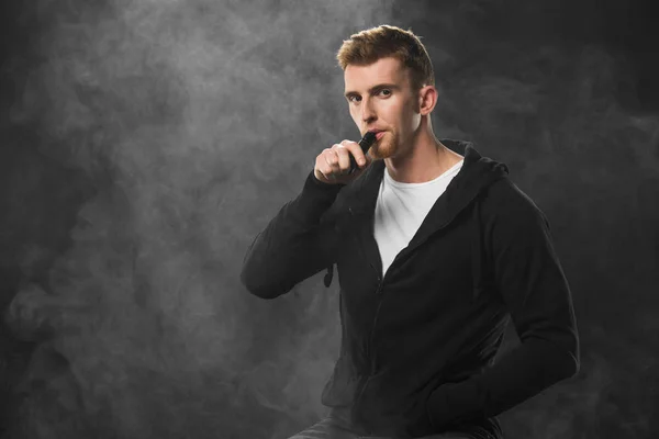 Joven barbudo vapeando cigarrillo electrónico rodeado de nubes de vapor - foto de stock