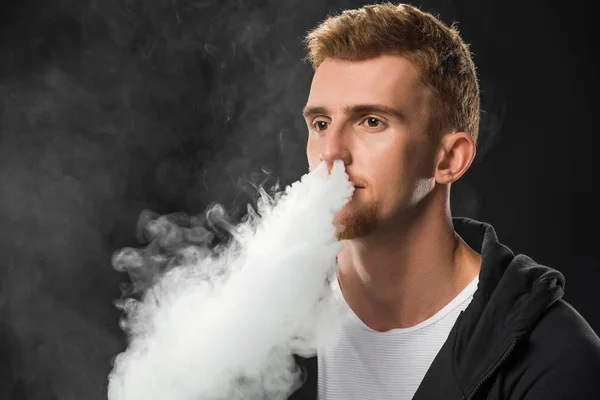 Jeune homme barbu expirant fumée de cigarette électronique entourée de nuages de vapeur — Photo de stock