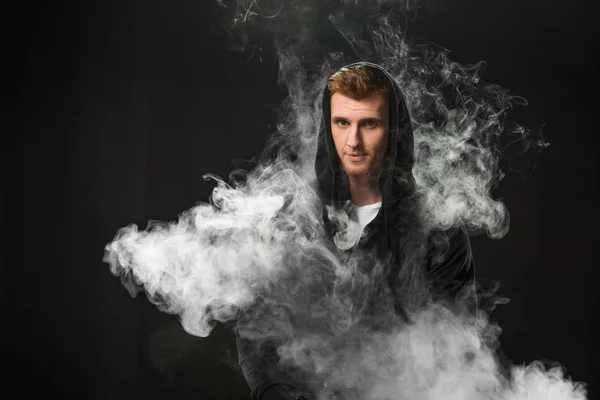 Jeune homme barbu vapotage cigarette électronique entourée de nuages de vapeur — Photo de stock