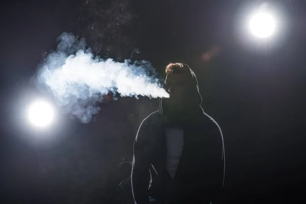 Joven barbudo exhalando humo de cigarrillo electrónico contra retroiluminación - foto de stock