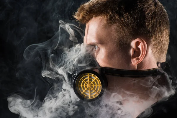 Man in black hood wearing protective filter mask surrounded by clouds of smoke — Stock Photo