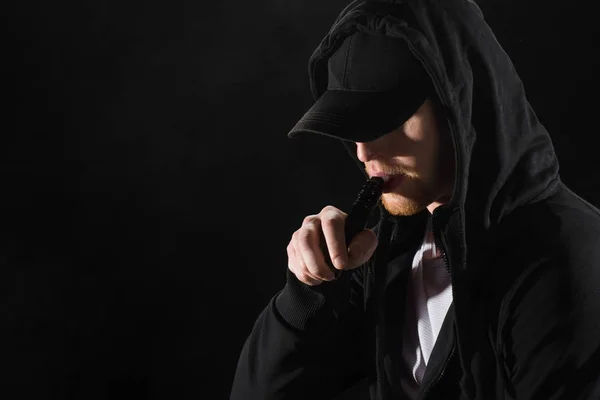 Junger bärtiger Mann mit Mütze, der elektronische Zigarette raucht, isoliert auf schwarz — Stockfoto