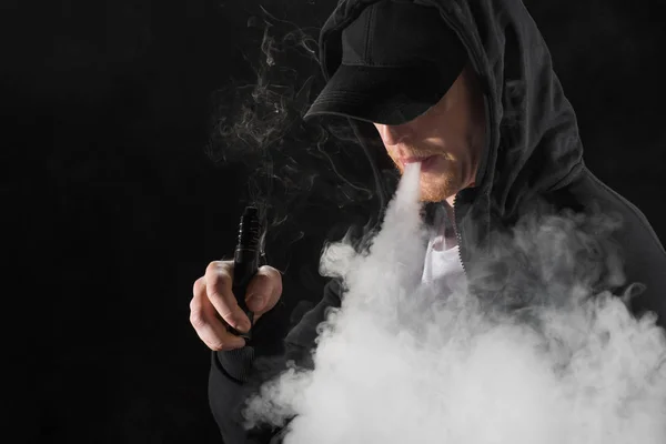 Молодой бородатый мужчина выдыхает дым электронной сигареты в окружении облаков пара — стоковое фото