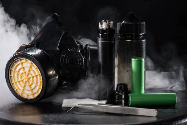 Electronic cigarette and protective filter mask with clouds of smoke on dark background — Stock Photo