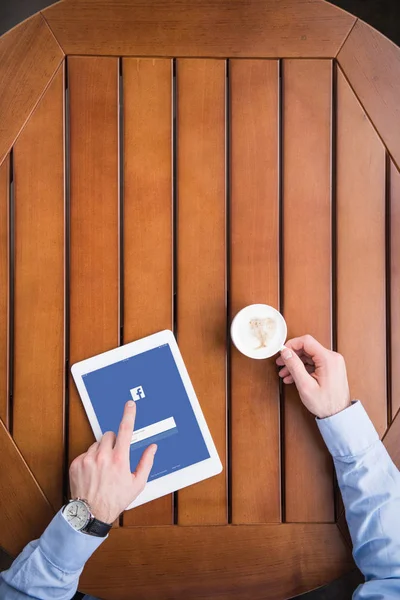Image recadrée de l'homme assis avec du café et chargé page facebook sur tablette — Photo de stock