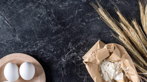 Top view of arrangement of raw eggs, flour and wheat on dark marble tabletop — Stock Photo