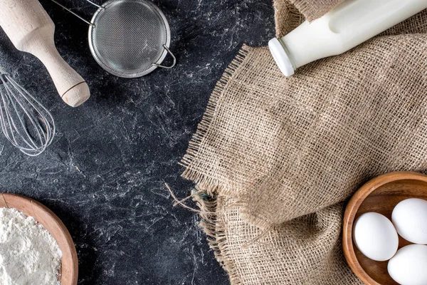 Flat lay with bottle of milk, flour, raw eggs and various supplies for bakery on dark tabletop — Stock Photo