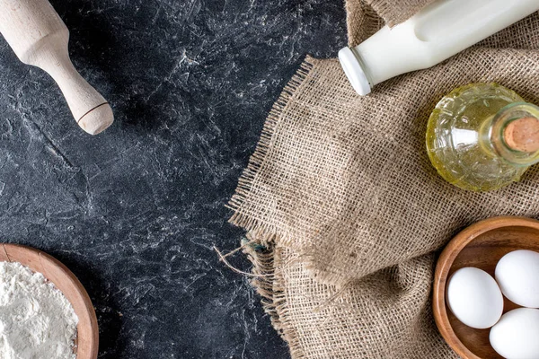 Top view of bottles of oil and milk, flour, raw eggs and rolling pin on dark marble surface — Stock Photo