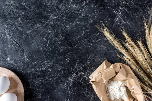 Top view of raw eggs, flour and wheat on dark marble surface — Stock Photo