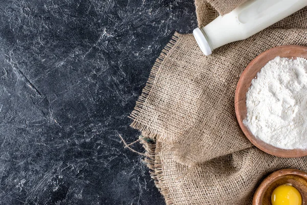 Draufsicht auf eine Flasche Milch, Eigelb und Mehl auf einem Sacktuch auf einer dunklen Marmortischplatte — Stockfoto