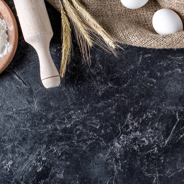 Flat lay with wheat, raw eggs on sack cloth and wooden rolling pin on dark marble surface — Stock Photo