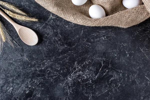 Flat lay with wheat, raw eggs on sack cloth and wooden spoon on dark marble surface — Stock Photo