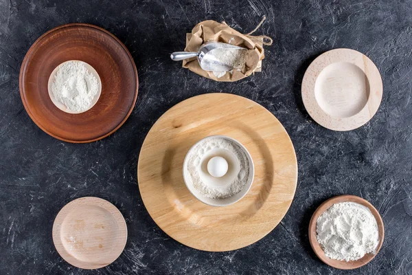 Flat lay with arrangement of wooden plates with flour and raw egg on dark marble tabletop — Stock Photo