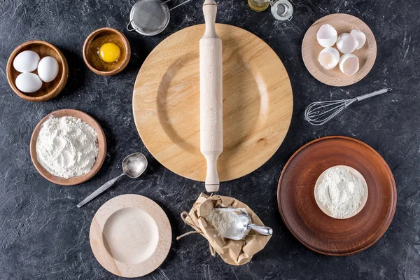 Flat lay with rolling pin, wooden plates and bakery ingredients on dark surface — Stock Photo