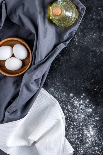 Vista dall'alto di uova crude in ciotola, bottiglia di olio e farina su superficie di marmo scuro — Foto stock