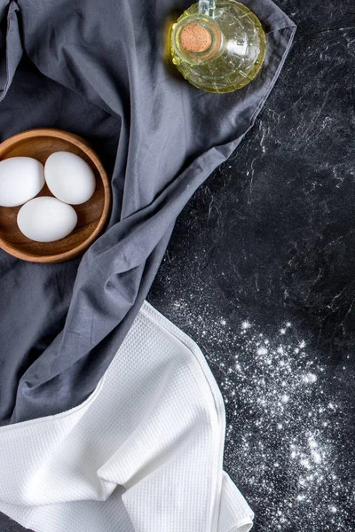 Top view of raw eggs in bowl, bottle of oil and flour on dark marble surface — Stock Photo