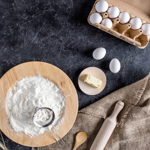 Vue du dessus des œufs crus, de la farine, du beurre et des ustensiles de cuisine pour la boulangerie sur table sombre — Photo de stock