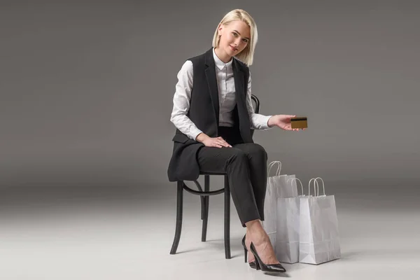 Mujer con estilo con tarjeta de crédito en la mano sentado en la silla cerca de bolsas de compras - foto de stock