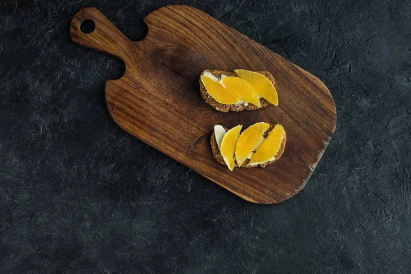 Vista superior de aperitivos con piezas de naranja para el desayuno en la tabla de cortar en la superficie oscura - foto de stock