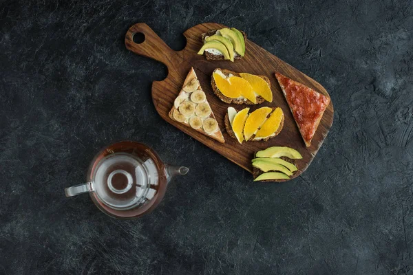 Flat lay with snacks for healthy breakfast and teapot on wooden cutting board on dark tabletop — Stock Photo