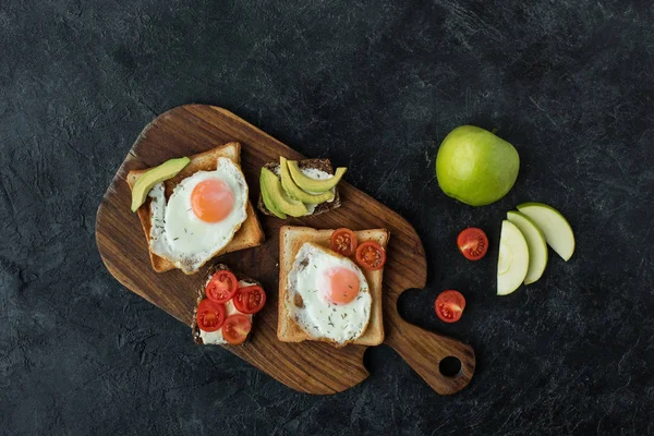 Vista superior de las tostadas con huevos fritos para el desayuno en la tabla de cortar de madera en la superficie oscura - foto de stock