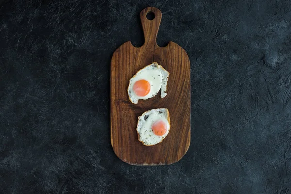 Top view of fried eggs on wooden cutting board on dark surface — Stock Photo