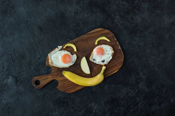 Vista superior de la cara sonriente hecha de comida para un desayuno saludable en la tabla de cortar - foto de stock