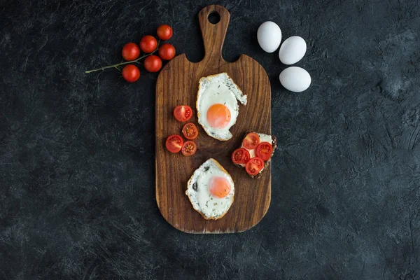 Top view of arrangement of fried eggs and cherry tomatoes on wooden cutting board on dark surface — Stock Photo