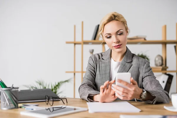 Focused businesswoman using smartphone while sitting at workplace in office — Stock Photo